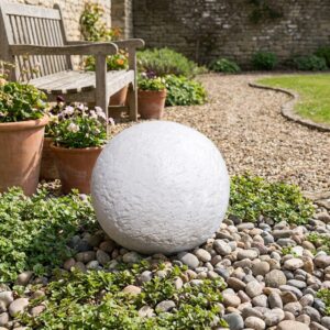 Boule décoration jardin extérieur en pierre blanche de 24 cm de diamètre reposant sur des galets, avec banc de jardin en arrière-plan. Référence du produit : 212064